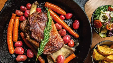  Cacerola de hierro con un corte de carne, romero, zanahorias y papas, acompañada de ensalada y papas fritas.
