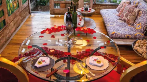 Vista superior de una mesa de cristal decorada con pétalos rojos, vajilla servida, copas y vino junto a un sofá.