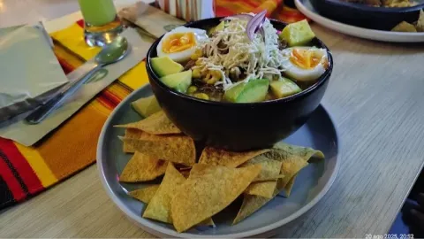Bowl de ensalada con aguacate, huevo cocido, queso rallado y otros ingredientes, acompañado de nachos crujientes.
