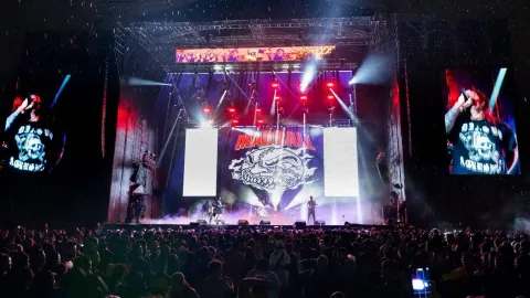 Escenario de concierto con banda de rock en vivo, luces intensas y gran público frente a la tarima durante un evento nocturno.