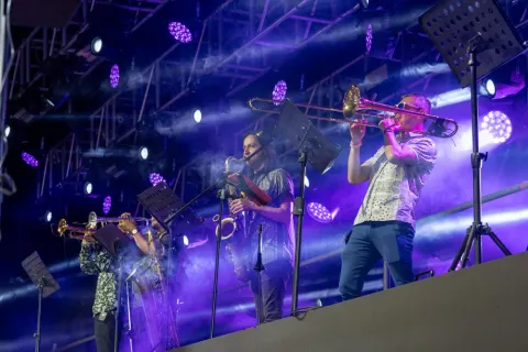 Músicos en escenario tocan trombón y saxofón bajo luces moradas durante un concierto en vivo.