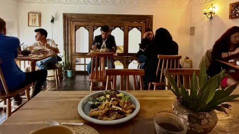 Salón acogedor de restaurante con comensales disfrutando sus platos, mesas de madera, decoración tradicional y detalles de vegetación.