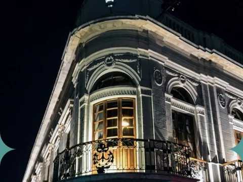 Fachada de un edificio histórico iluminado de noche, con ventanales de madera, balcones de hierro forjado y ornamentación clásica.
