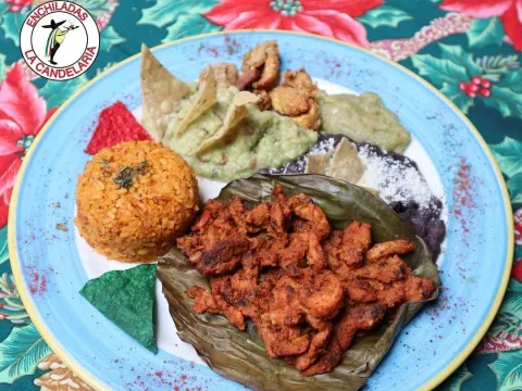 Plato de comida mexicana con pollo sazonado, arroz, guacamole, frijoles, totopos y salsas, servido sobre hoja de plátano.