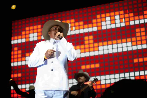 Cantante con traje blanco y sombrero interpreta con micrófono frente a una pantalla luminosa en concierto nocturno.