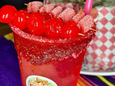 Bebida roja decorada con frutas, dulces y un borde cubierto de chile.