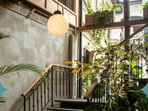 Escalera interior rodeada de vegetación, con barandas de hierro forjado, detalles en madera y una lámpara de luz cálida.