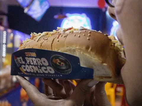 Perro caliente de El Perro Cósmico servido con pan de ajonjolí y abundantes toppings.