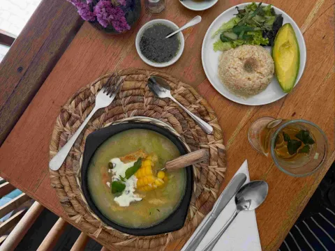  Plato vegetariano como ajiaco con sopa de vegetales, maíz, crema y hierbas, acompañado de ensalada, aguacate y una bebida caliente.