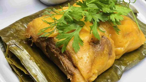 Preparación tradicional servida sobre hoja de plátano, con una cubierta dorada y suave, acompañada de hierbas frescas.