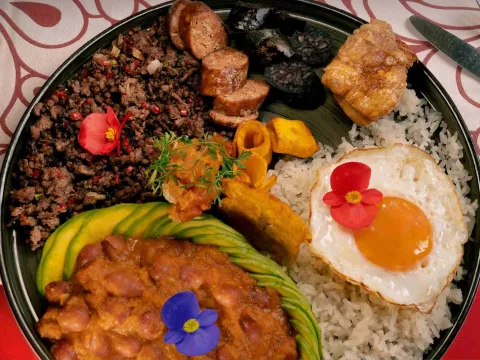  Plato típico colombiano con arroz, fríjoles, carne molida, huevo frito, aguacate, chorizo, plátano y chicharrón, presentado con flores comestibles.