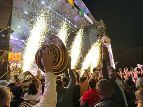 Público celebra frente a un escenario con fuegos artificiales, levantando sombreros durante un concierto nocturno.