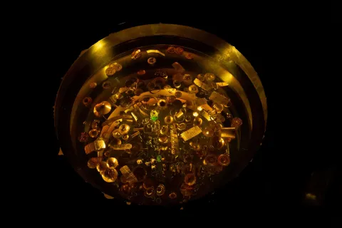 Una pieza circular de oro exhibe numerosas joyas y objetos de orfebrería precolombina, resaltados por una iluminación tenue.