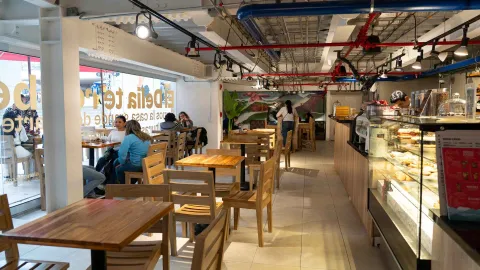 Comedor interior con mesas de madera, ventanal hacia la calle, vitrina de repostería y tuberías industriales.
