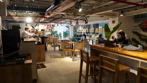 Vista del salón con barra alta de madera, personas trabajando con computadores y baristas al fondo preparando café. 