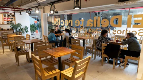 Salón con múltiples mesas de madera luminosas junto a ventanales con vinilos decorativos y clientes conversando. 