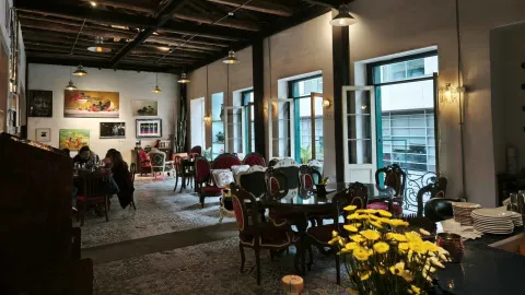 Salón de época con sillas de estilo clásico, grandes ventanales, pinturas en las paredes y flores amarillas. 