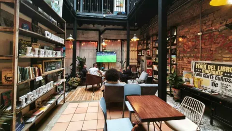 Patio interior con zona de librería, estantes con libros, mesas de madera y clientes viendo una televisión. 