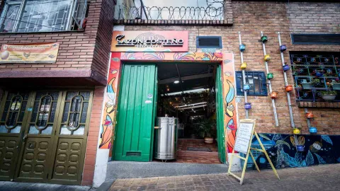 Fachada del restaurante Sazón Costeño con puerta verde abierta, barril ahumador y macetas coloridas en la pared.