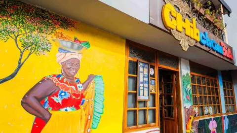 Plano medio del mural de la fachada con una mujer palenquera sonriendo sobre un fondo amarillo vibrante.