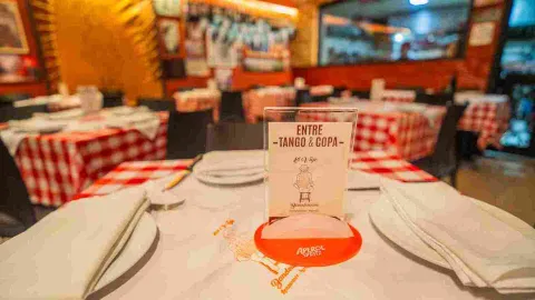 Mesa servida con mantel a cuadros rojos y blancos, servilletas y cartel promocional de Entre Tango & Copa.