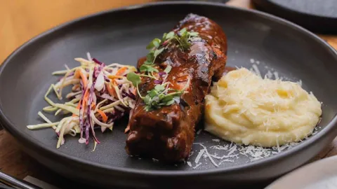 Plato servido con costilla bañada en salsa barbacoa, acompañado de puré de papa y ensalada de col. 