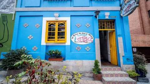 Puerta amarilla abierta con reja en un edificio azul. Se observan escalones de azulejos, un letrero y el número 6-17.