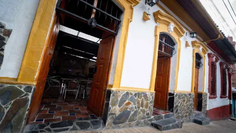 Perspectiva en ángulo de fachada colonial blanca con marcos amarillos, zócalos de piedra y puertas de madera abiertas.