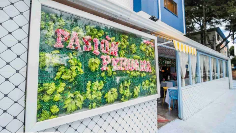 Muro exterior con azulejos geométricos y gran letrero del restaurante elaborado con flores rosadas sobre follaje verde. 