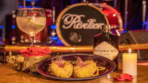 Un cóctel de ginebra y Hendrick's, junto a dos aperitivos empanizados, una vela y pétalos de rosa, con una batería de fondo que dice "Rebellion".