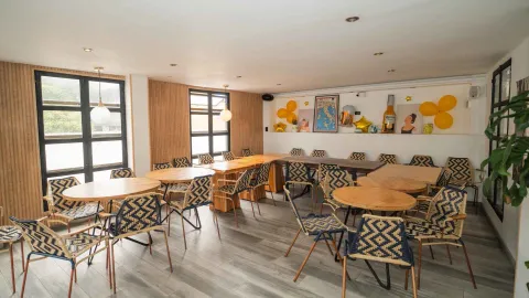 Salón comedor del restaurante con mesas de madera claras, sillas tejidas con patrones geométricos en azul y blanco, y globos amarillos de decoración.