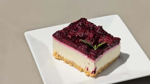 Porción cuadrada de cheesecake con mermelada de frutos rojos y una hoja verde servida en plato cuadrado blanco. 