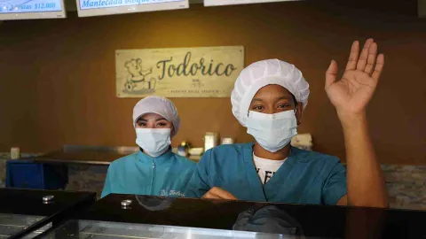 Dos empleadas con tapabocas y cofia azul detrás del mostrador, una de ellas saludando con la mano levantada. 