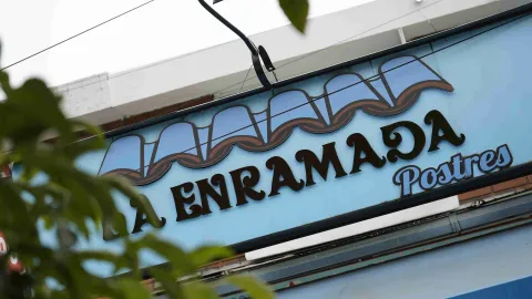 Letrero exterior azul de La Enramada Postres visto entre ramas de árboles. 