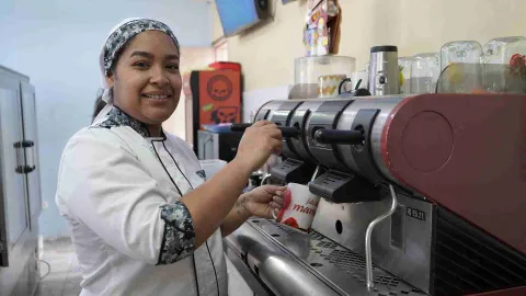 Una chef sonríe mientras maneja una máquina de café expreso profesional, sosteniendo una taza decorada. 