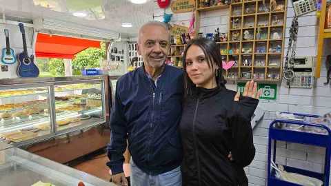  Un hombre mayor con chaqueta azul y una mujer joven con buzo negro posan sonriendo junto a la vitrina de postres. 