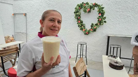 Mujer con filipina de cocina sostiene un vaso transparente con malteada frente a una pared con corazón floral. 