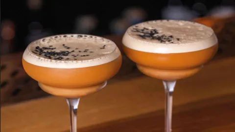 Dos copas de cóctel de tono ámbar con espuma y semillas de ajonjolí negro decorando la parte superior. 