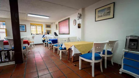 Sala de comedor con mesas vestidas con manteles amarillos y azules. Sillas blancas de madera y suelo de baldosas rojas.