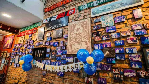 Plano detallado de pared de ladrillo rústico cubierta de retratos vintage, cuchillos exhibidos y globos con pancarta de Feliz Cumpleaños.