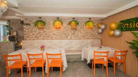 Comedor con sillas de plástico naranja, mesas con manteles blancos, pared de piedra y lámparas con follaje vegetal.