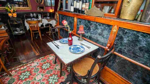 Mesa pequeña de mármol para dos personas con platos decorados, una flor roja y sillas de madera sobre piso hidráulico.
