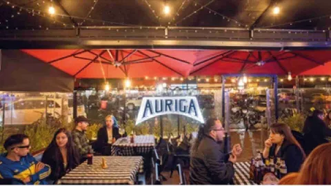 Clientes compartiendo en las mesas de la terraza exterior bajo sombrillas rojas con el letrero de Auriga al fondo. 