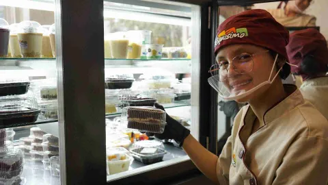 Pastelera sosteniendo una porción de torta empaquetada frente a una vitrina refrigerada llena de postres. 