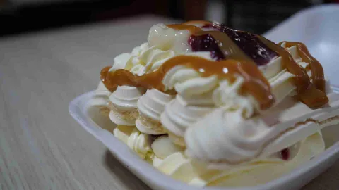 Postre de merengue con crema, decorado con salsa de mora y líneas de arequipe en recipiente blanco. 