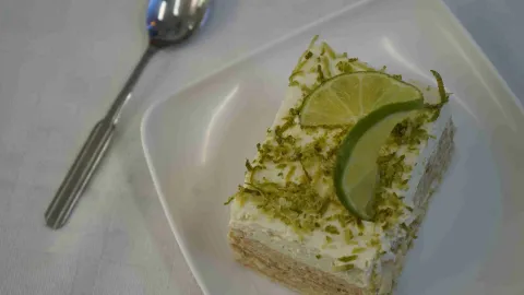 Porción cuadrada de postre de limón con ralladura y rodajas de lima sobre plato blanco. 