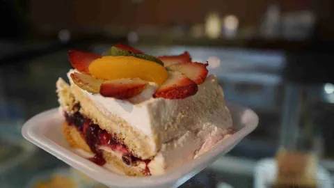 Porción de torta de crema con relleno de mermelada y topping de fresas, melocotón y kiwi en recipiente blanco. 