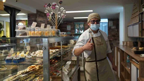 Pastelero con tapabocas y delantal haciendo la señal de aprobación junto a la vitrina con postres y ponqués. 