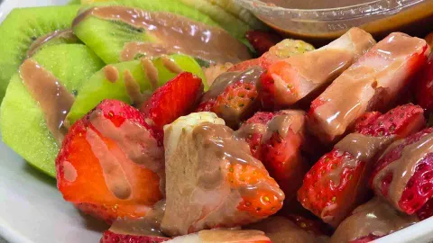 Un primer plano de trozos de kiwi y fresa fresca, bañados con salsa de chocolate sobre un plato blanco. 