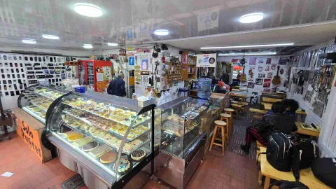Vista panorámica del interior de la pastelería con vitrinas exhibidoras llenas de postres, barra con bancos y clientes. 